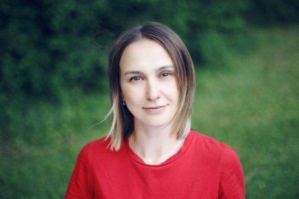 a young woman with brown shoulder length hair and dark eyes is looking at the camera with a slight smile, in front of a green nature background