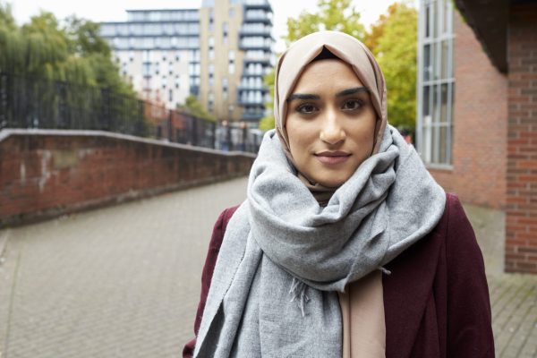 Young lady wearing a pale pink hijab and a grey scarf. She is looking into the camera with a slight smirk. She is standing in front of a brick wall and an apartment building in the background.