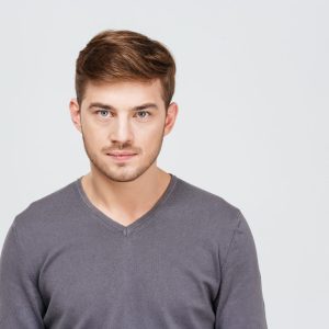 A young man with short brown hair, wearing a grey jumper, is looking straight ahead at the camera with a calm expression.