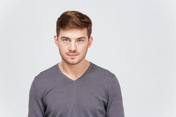 A young man with short brown hair, wearing a grey jumper, is looking straight ahead at the camera with a calm expression.